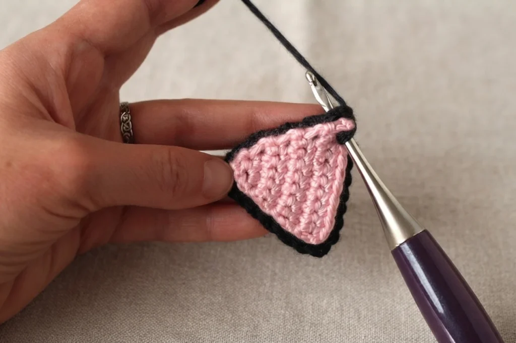 A hand holding a small pink crocheted triangle with a black crocheted edge, a crochet hook inserted through the top corner, against a plain beige fabric background.