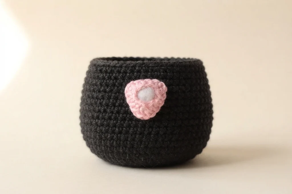 Black crocheted amigurumi body in progress with a small pink crocheted nose piece attached to the front, resting on a plain beige surface.