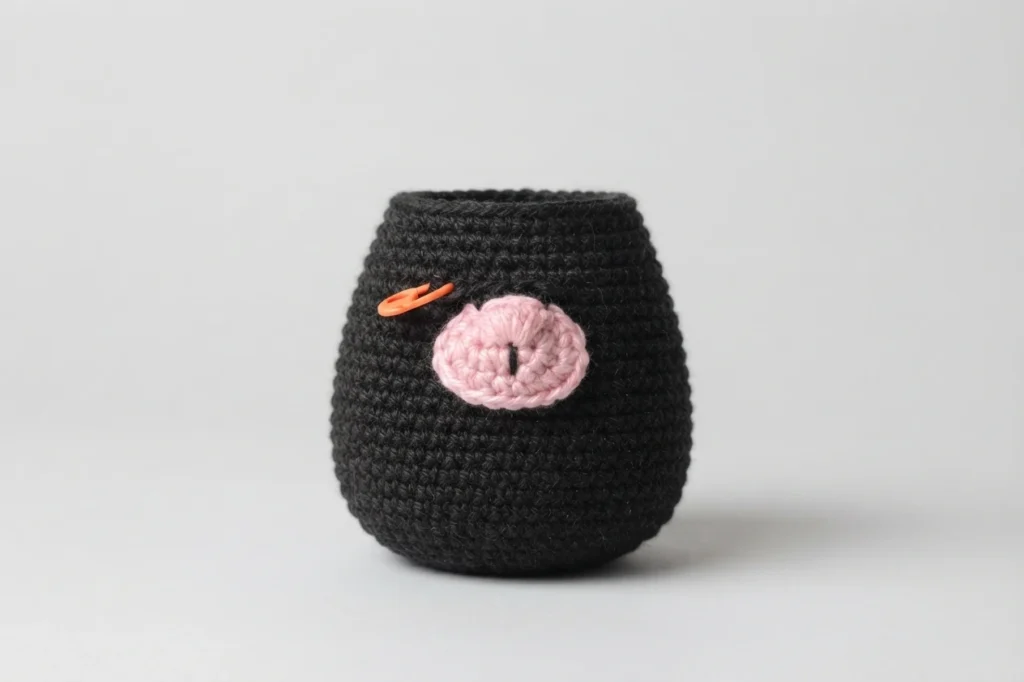 Dark charcoal crocheted gnome body with a pink crocheted nose piece attached to the front and an orange stitch marker clipped near it, on a plain gray background.