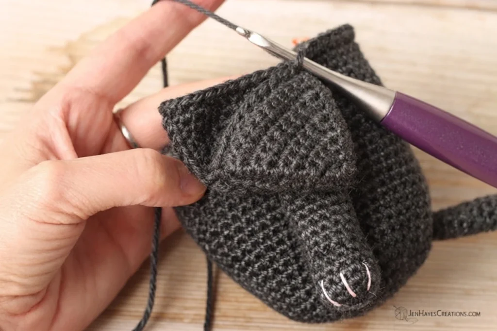 Close-up of hands holding a dark gray crocheted gnome body while a purple crochet hook joins a crocheted ear to it, with pink stitch markers marking the muzzle area and loose yarn ends hanging down.