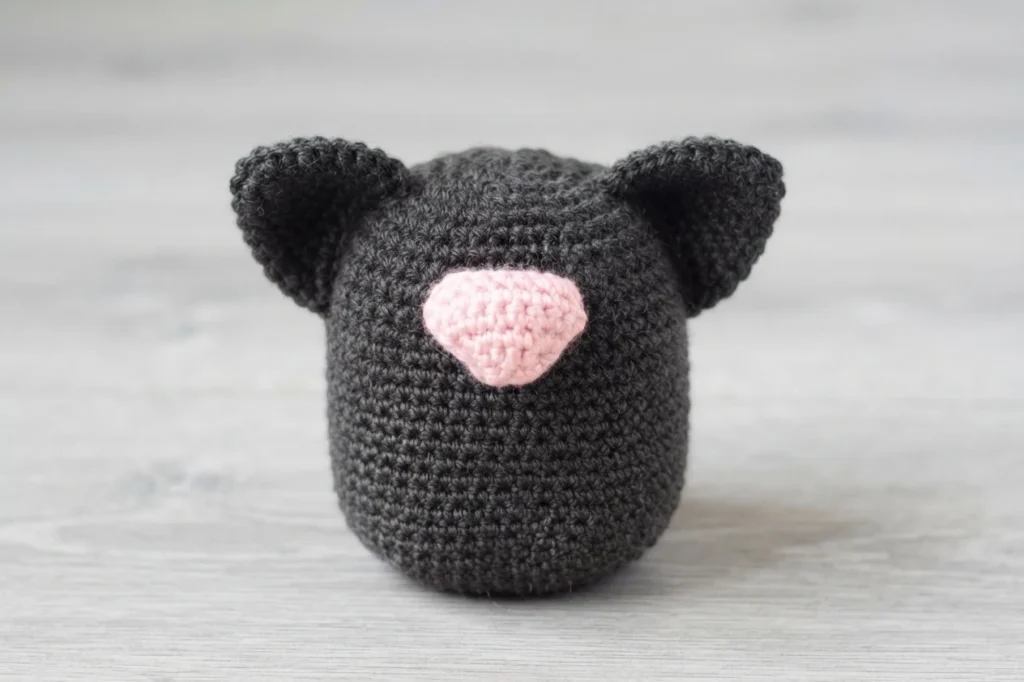 Corps de gnome chat au crochet en fil gris anthracite, deux oreilles jointes de chaque côté du haut et un museau rose en forme de coeur, posé sur une surface en bois grisé.