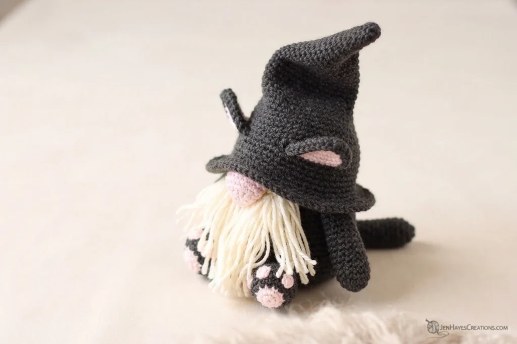A crochet cat gnome in charcoal gray yarn with a tall pointed hat, pink lined cat ears, a pink nose, a cream fringe beard and paws with pink pads, photographed from the side against a plain cream background with a soft textured mat.