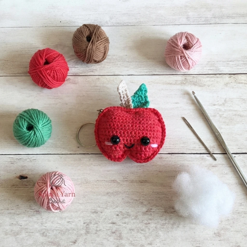 Crocheted red apple keychain with black safety eyes, pink cheeks and a small stitched smile, a cream stem and a green leaf, lying on a white wood surface beside balls of red, brown, green and pink yarn, a metal crochet hook, a tapestry need