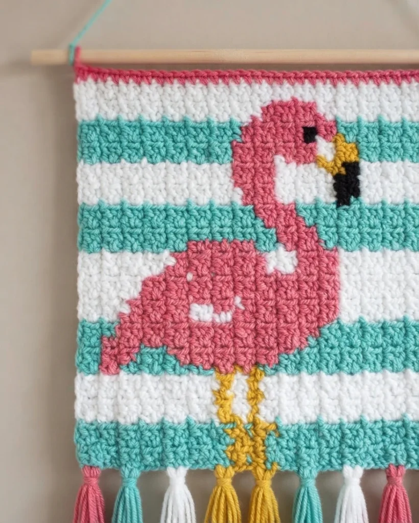 Crocheted corner to corner wallhanging hanging from a wooden dowel, showing a pink flamingo with a yellow and black-tipped beak and yellow legs against alternating teal and white stripes, finished with tassels along the bottom edge.