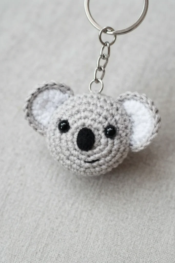 Crocheted grey koala head keychain with white inner ears, black safety eyes and a black oval nose on a metal chain and ring, lying on light fabric