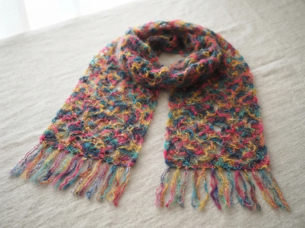 Multicolored variegated mohair lace scarf with fringed ends, folded loosely on a light linen surface near a window.