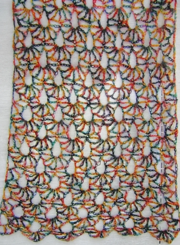 Close view of a crocheted openwork fabric in variegated orange, green, red and blue yarn, showing repeating fan and diamond shaped lace motifs with large open holes against a plain white background.