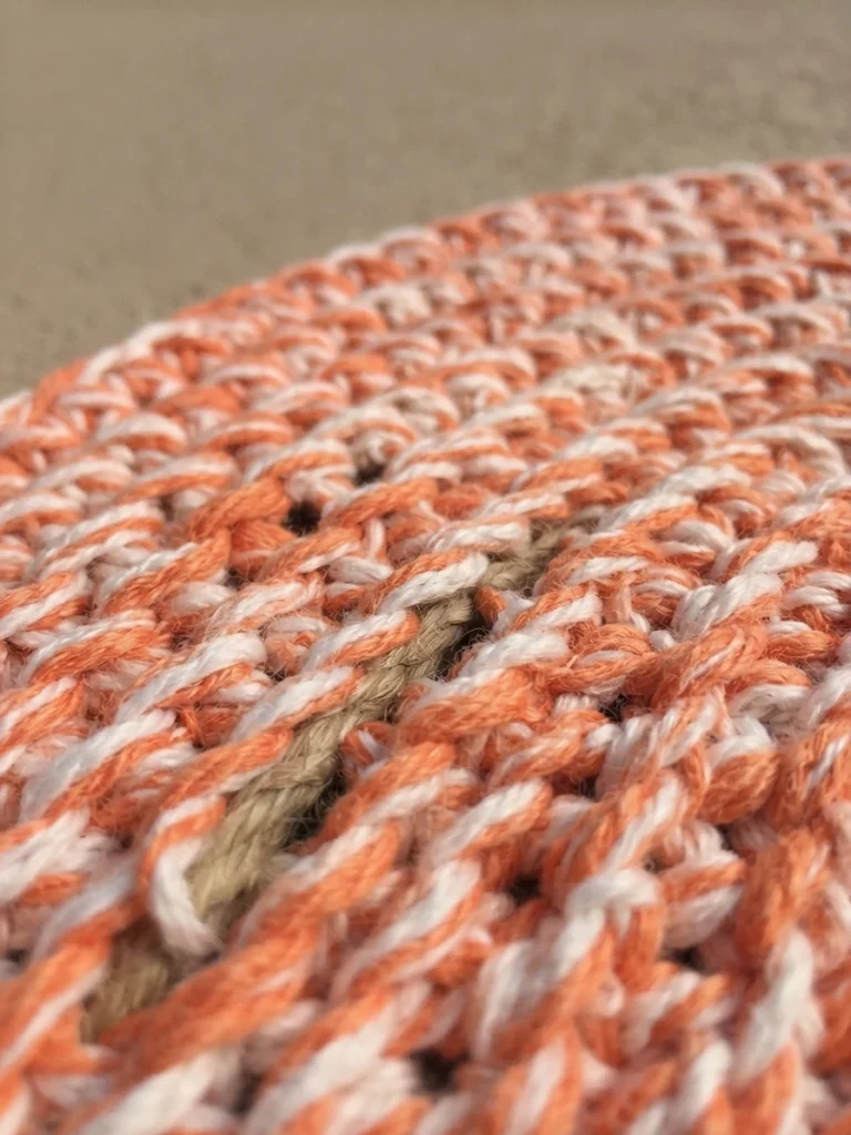 Close-up of a round crocheted scrap yarn rope bath mat in white and coral-orange yarn with the rope core visible between the stitches.