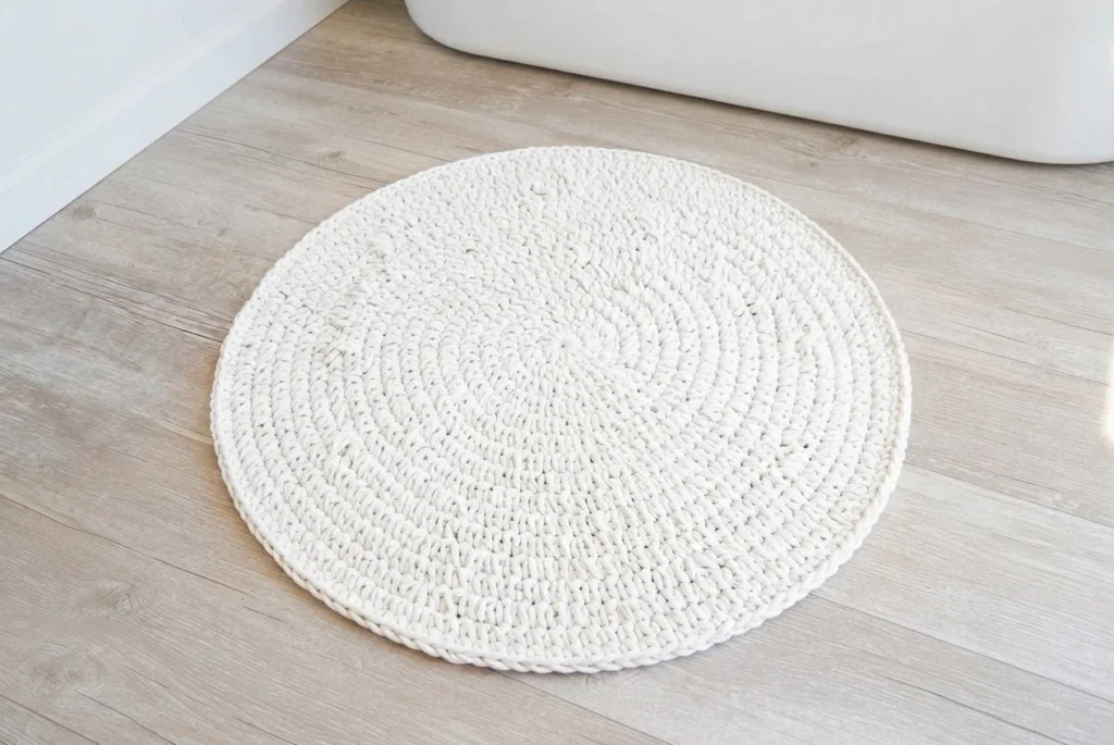 Round white crocheted bath mat on a light wooden floor near a bathtub