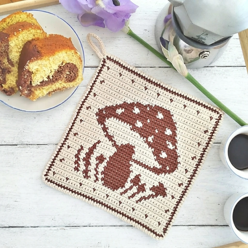 Square crocheted potholder in cream with a brown mushroom motif and a dotted border, hanging by a loop, styled on a white wooden table with sliced cake, coffee cups and a purple flower.