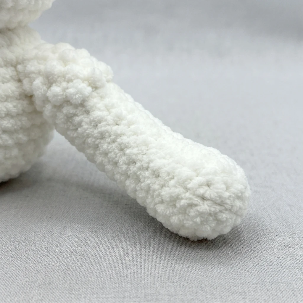 Close-up of a white plush crocheted arm attached to a body, resting on light grey fabric