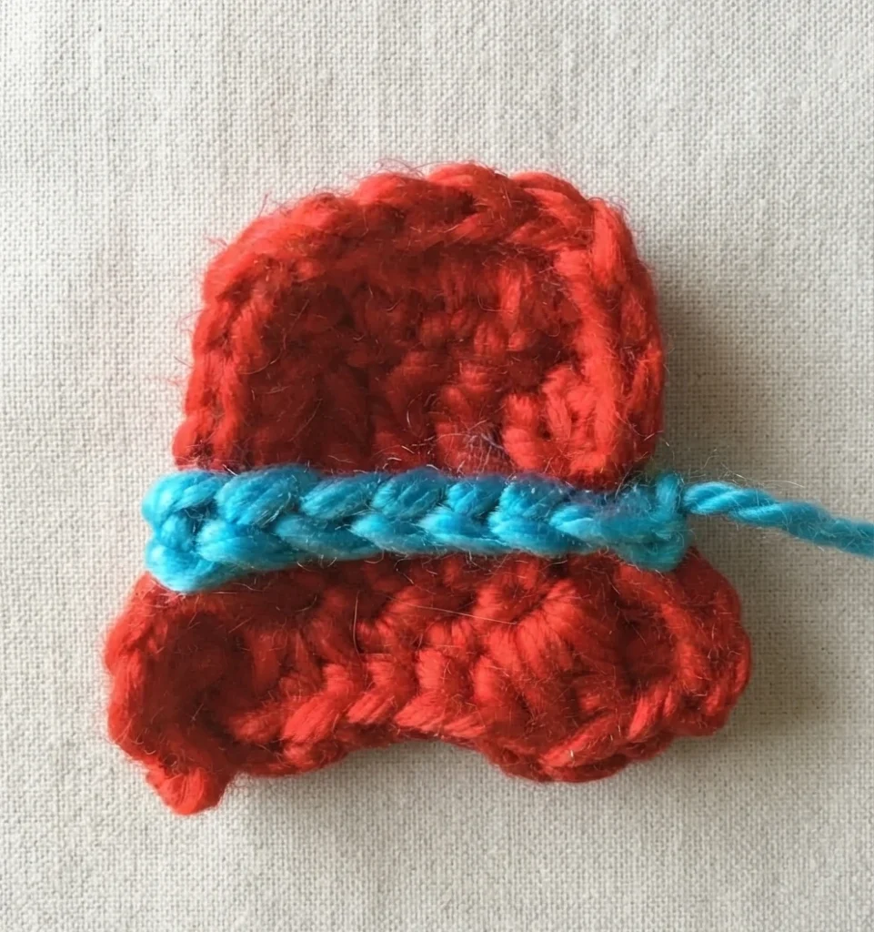 Crochet top hat in red yarn with a blue chain trim worked across the front, shown on a light beige fabric surface with a loose blue yarn end extending to the right