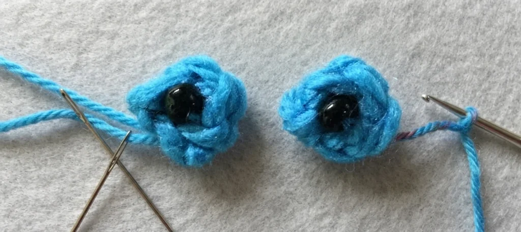 Two small blue crocheted eye pieces with black beads at their centres, resting on a light grey textured surface, with a yarn needle and blue yarn on the left and a crochet hook with blue yarn on the right.