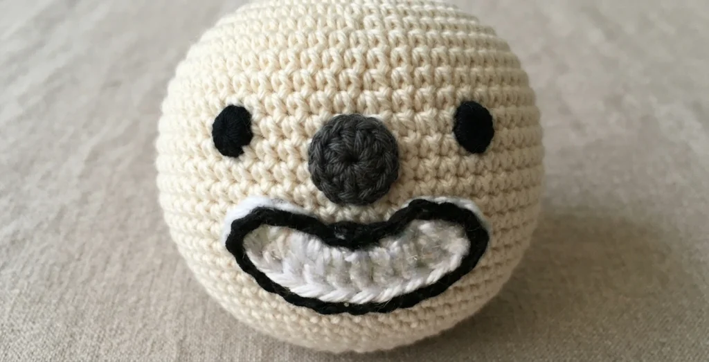 Crochet clown head in cream yarn with two black embroidered dot eyes, a grey crocheted nose and a white crocheted mouth piece outlined in black slip stitch, resting on a beige woven surface.