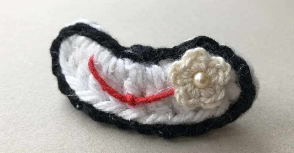 Crochet clown mouth piece in white with a black slip stitch border and a red embroidered smile, with a cream crocheted flower and a pearl bead attached at one side, photographed on a plain light background.