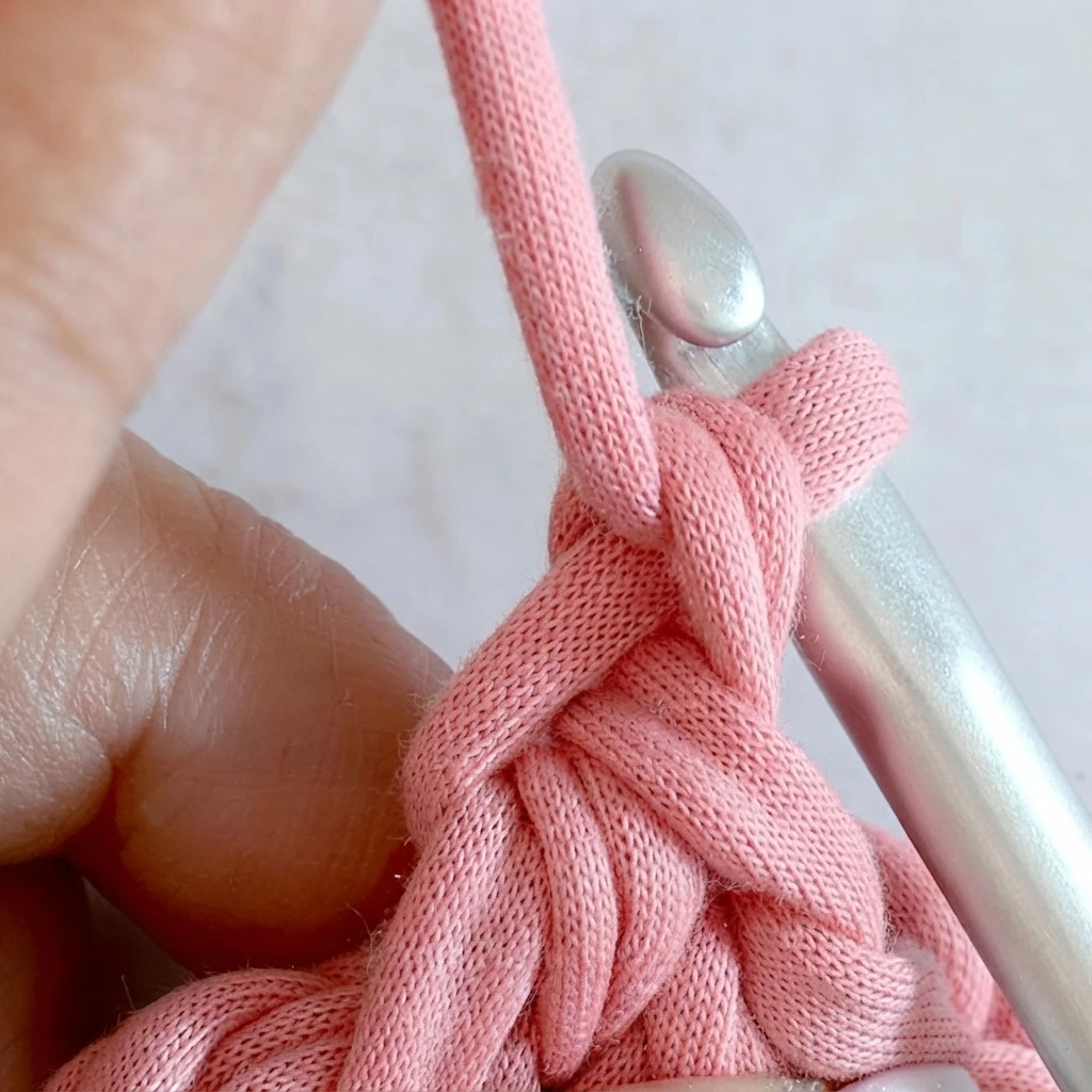 Close-up of pink t-shirt yarn stitches on a silver crochet hook held by fingers, showing an in-progress crochet heart step