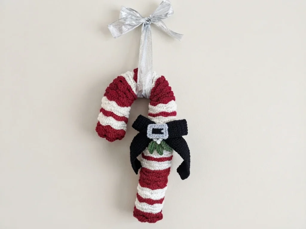 Crocheted candy cane wreath with burgundy and white stripes, a black bow with a silver buckle, small green mistletoe leaves, and a silver ribbon hanging loop at the top, photographed against a plain light background.