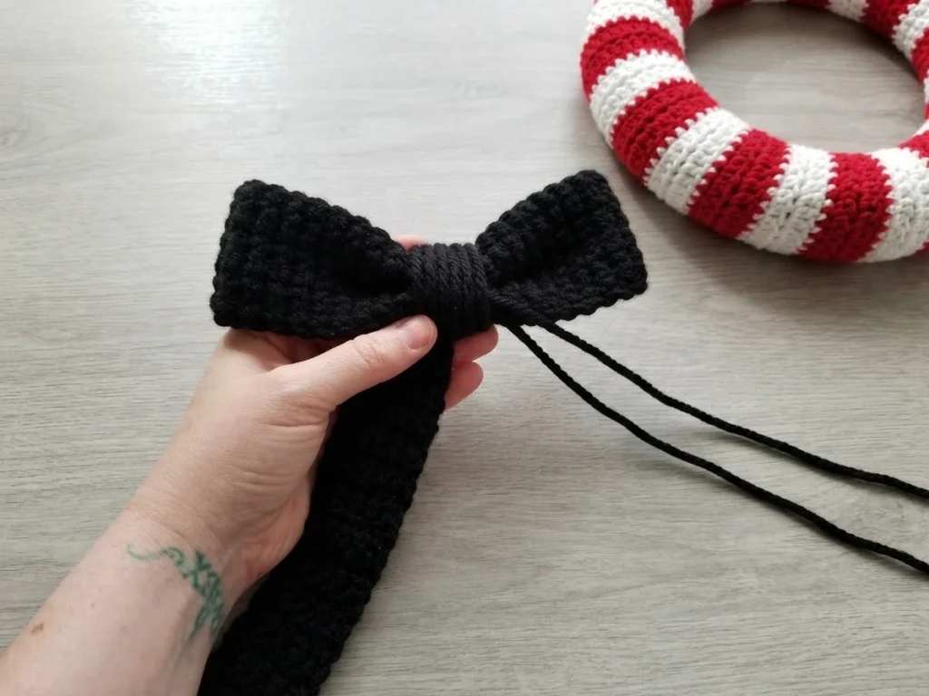 Hand holding a folded black crocheted bow with a wrapped center and two long black yarn tails, a red and white striped crochet wreath resting on a light wood surface in the background