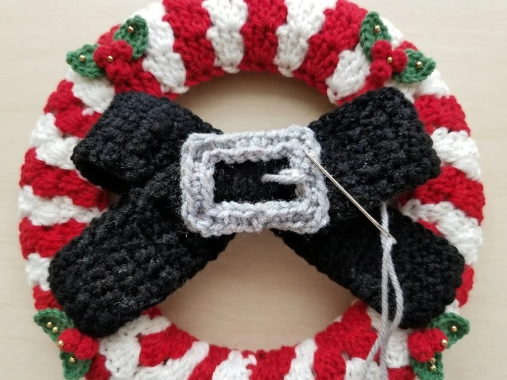 Close-up of a black crochet bow with a light gray crochet buckle on a red and white striped wreath, a tapestry needle and gray yarn mid-stitch through the buckle, with small green leaves and gold beads around the ring.