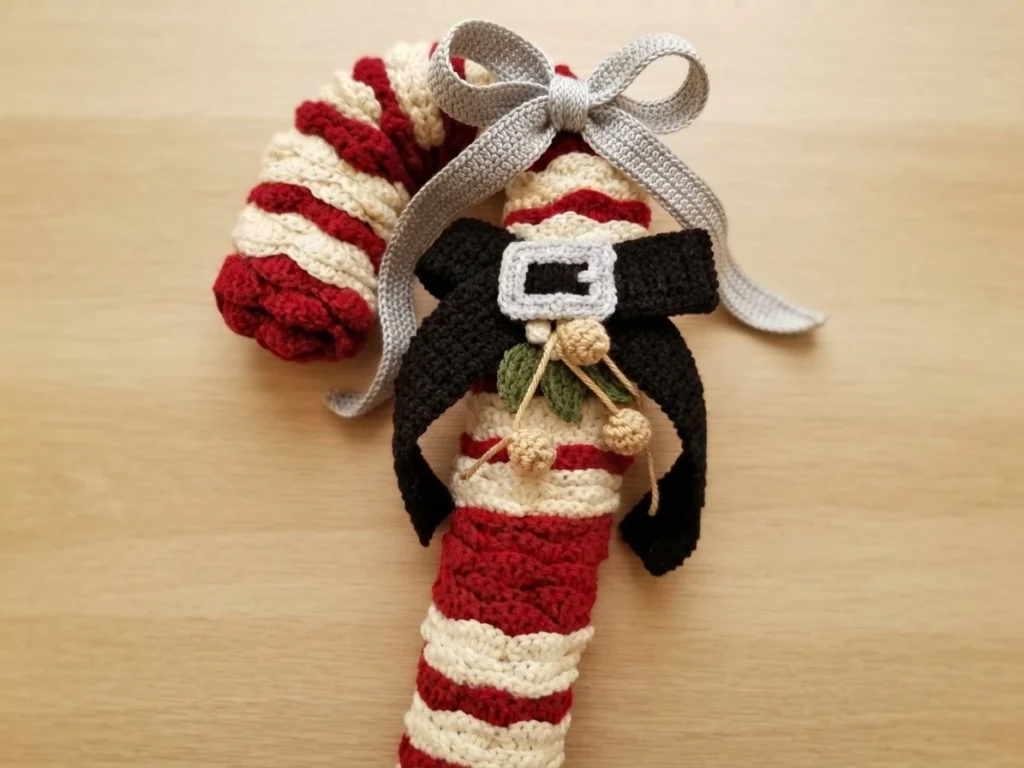 Crochet candy cane wreath in red and white stripes with a black bow, silver buckle and a mistletoe sprig with berries, hung from a silver ribbon bow on a light wood floor.