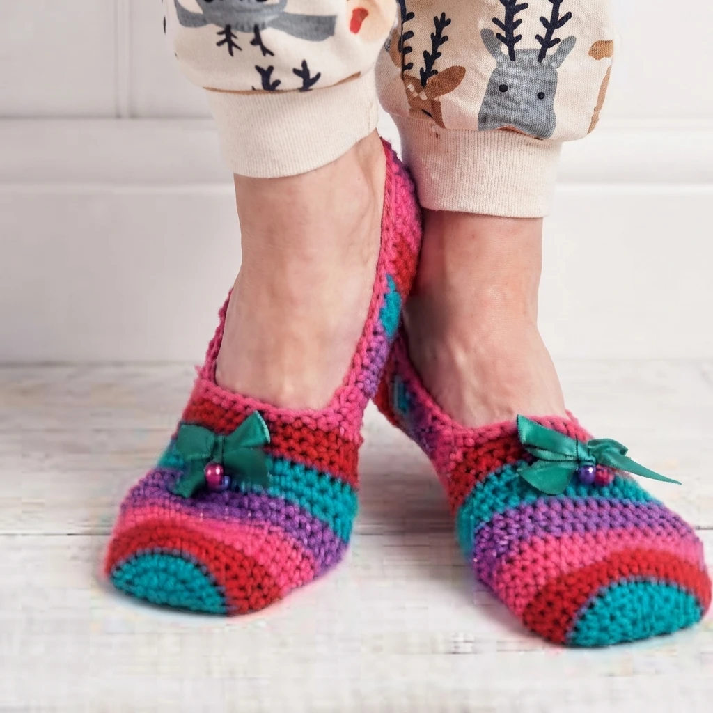 Pair of crocheted slippers in pink, red, purple and teal stripes with green ribbon bows and small beads, worn on bare feet with patterned cream pyjama trousers, standing on a white painted wooden floor.