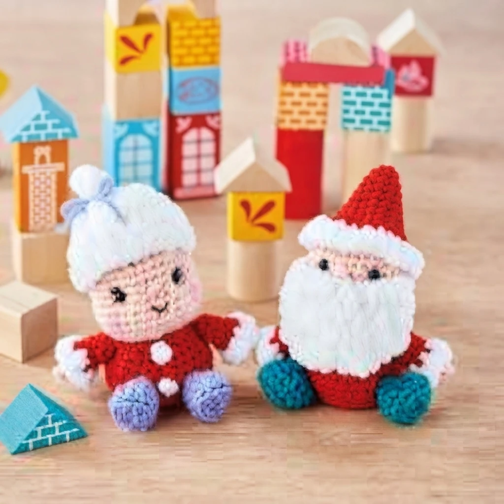 Two crochet Christmas dolls on a light wooden floor, Mrs Claus with a white bonnet, blue bow and lavender bootees beside Santa with a red hat, white beard and turquoise bootees, surrounded by colourful wooden toy blocks.