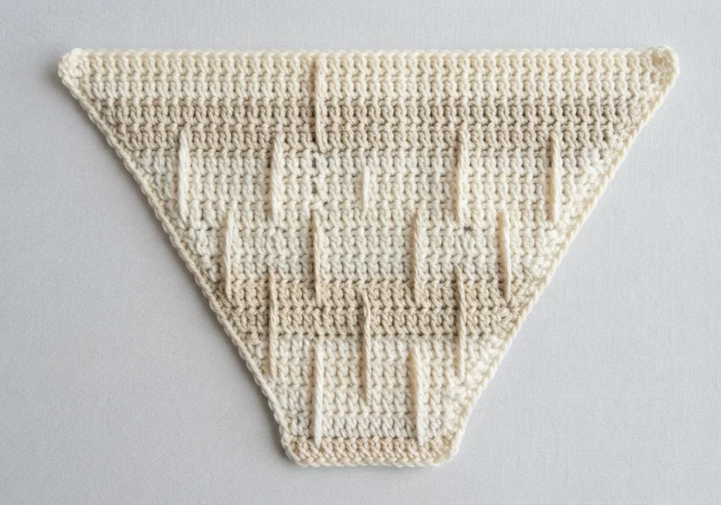 Triangular cream crocheted piece with light beige shading and raised vertical spike stitches, laid flat on a pale background