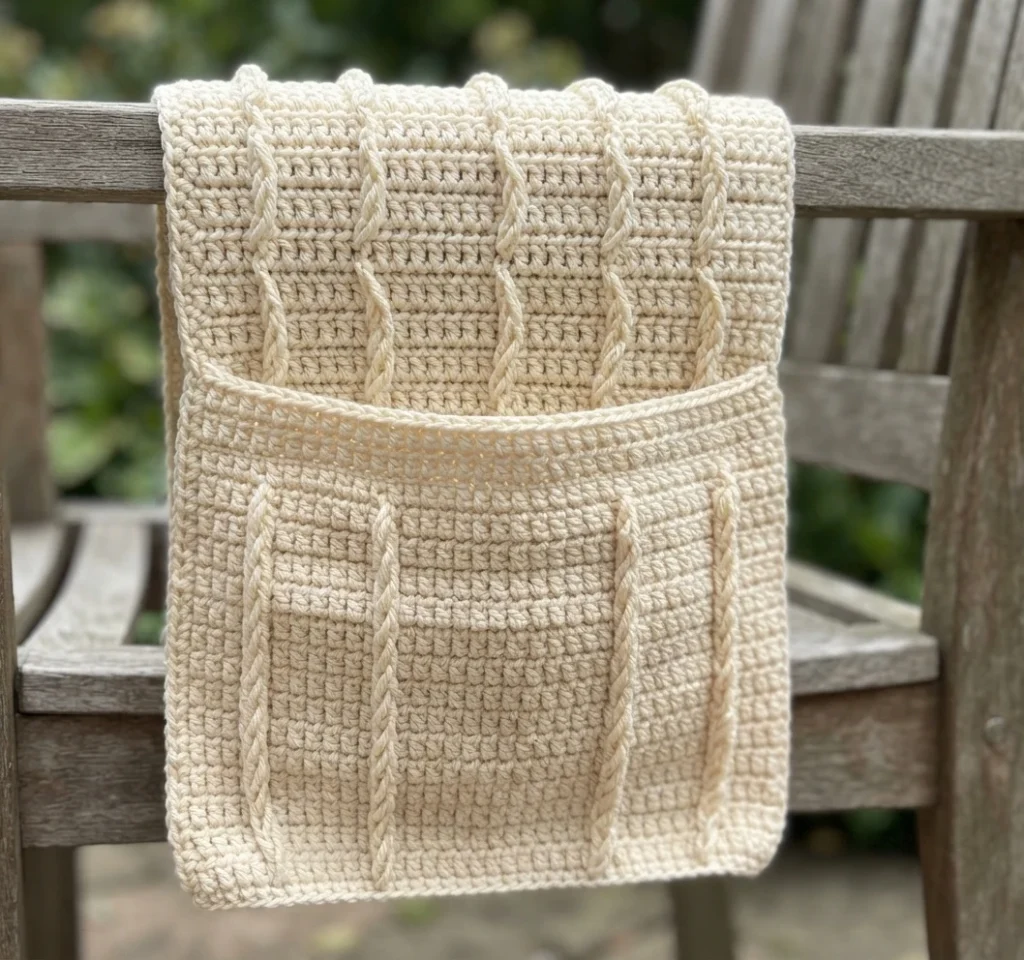 Cream crochet chair caddy with a front pocket and vertical raised stitch lines draped over a wooden chair arm outdoors