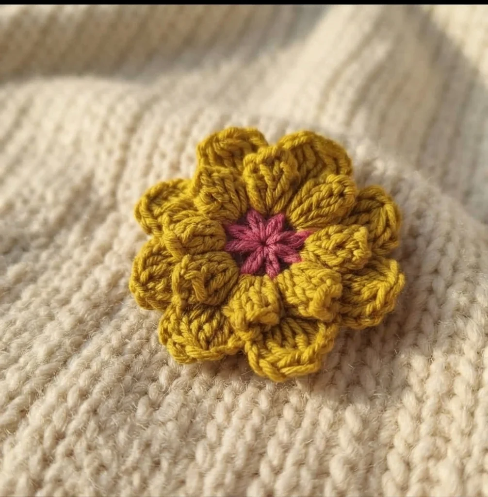 A mustard yellow crocheted flower with a pink center pinned on cream knitted fabric