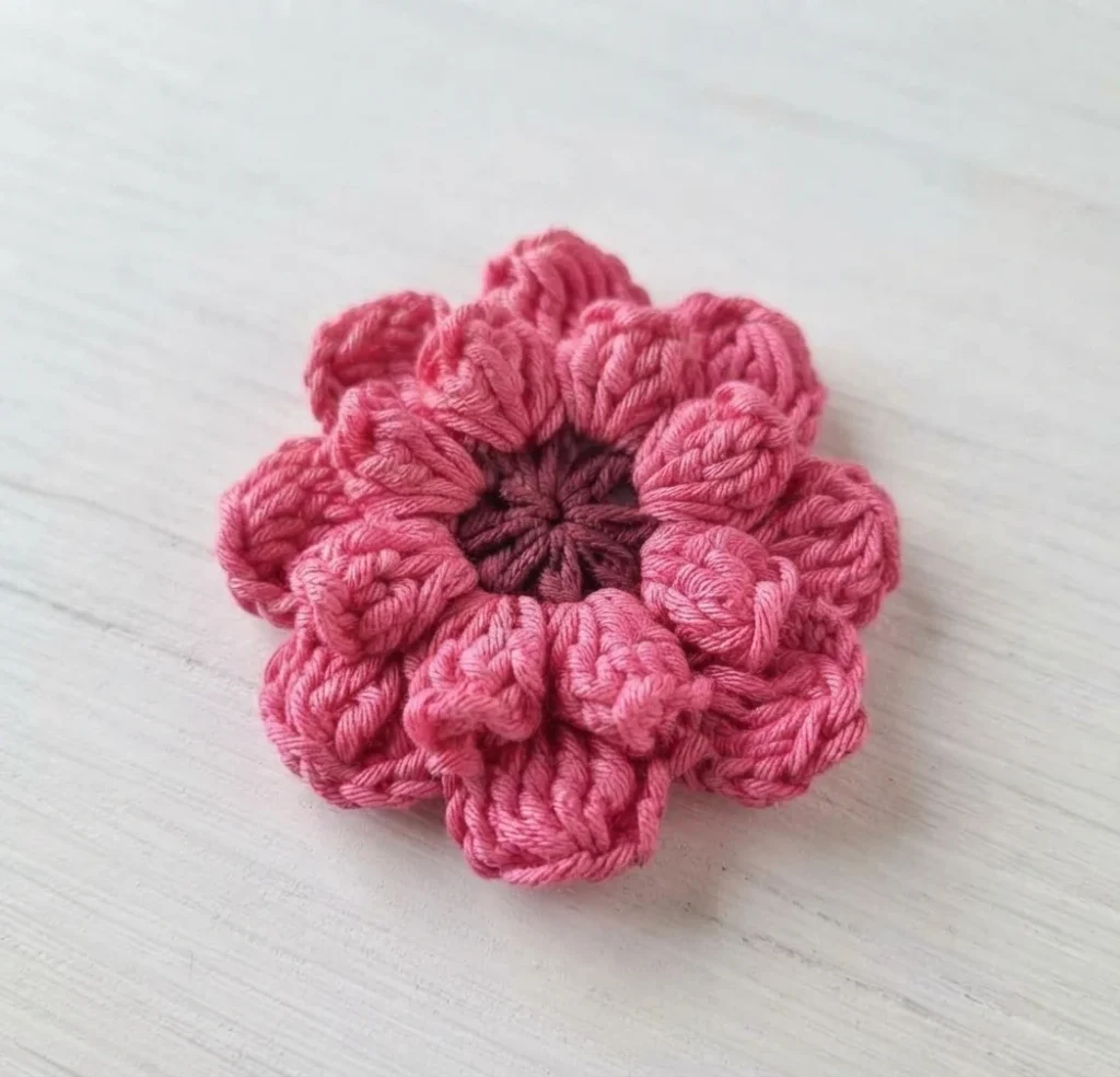 A single pink crocheted flower with a dark plum center resting on a light wood surface.