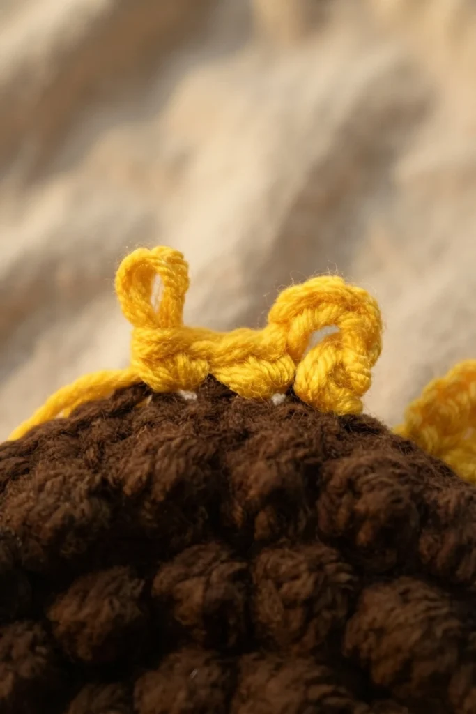 Close-up of two yellow crochet loops beginning to form petals on a textured brown crocheted sunflower center.