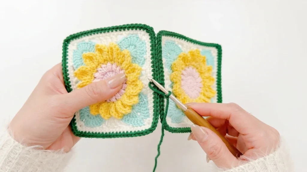 Hands holding two granny squares with yellow flowers and pink centers while slip stitching their green edges together with a crochet hook