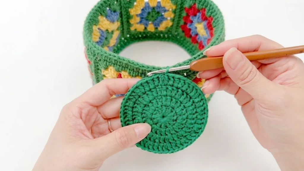 Hands holding a green crocheted circular bag bottom with a wooden handled crochet hook, joining it to a colorful granny square bag body