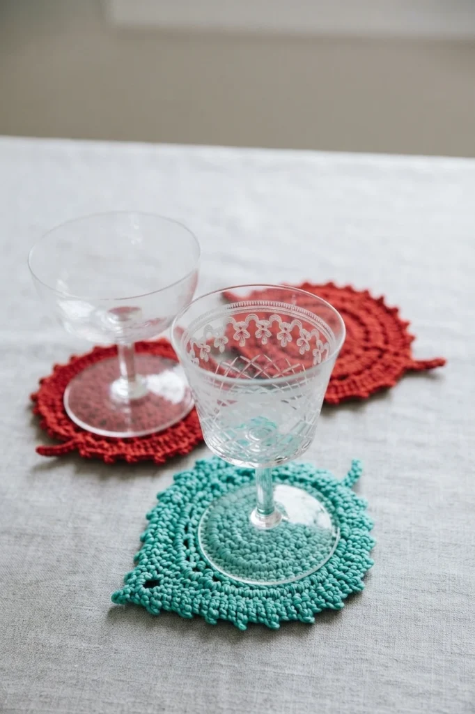 Two clear stemmed glass coupes resting on crocheted leaf-shaped coasters, one teal in front and one red behind, arranged on a light linen tablecloth.