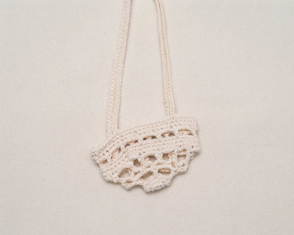 Cream crochet plant hanger with openwork body and long hanging cords laid flat on a light plain fabric background