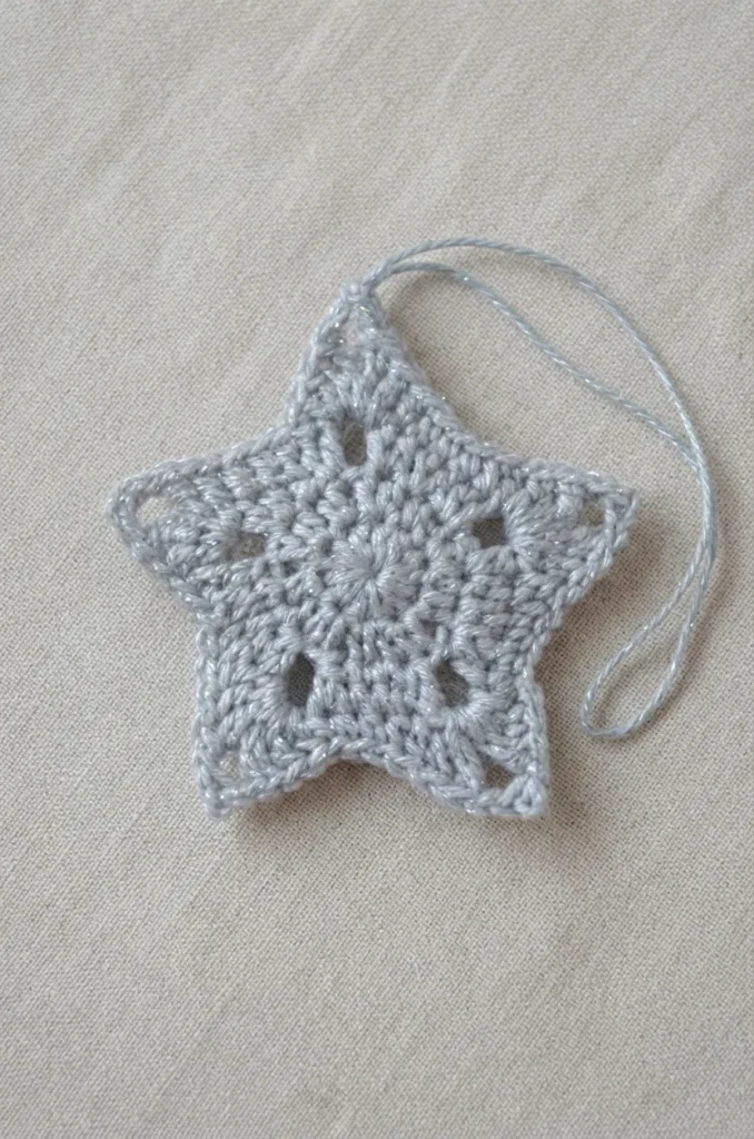 Étoile au crochet argentée et brillante avec une boucle de suspension, posée sur du tissu clair.