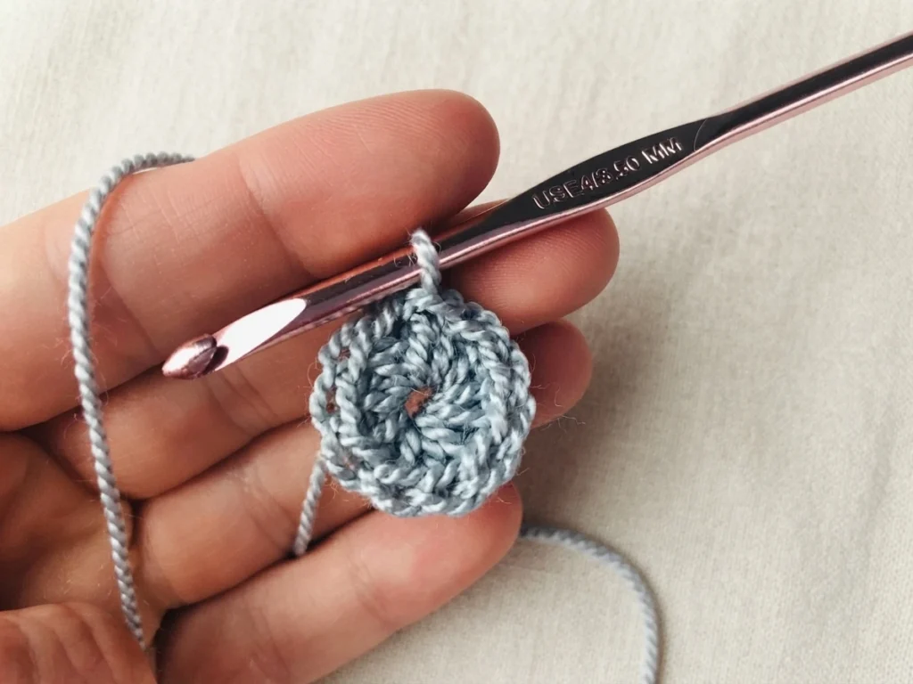 A hand holds a small gray crocheted circle on a hook with the yarn attached.