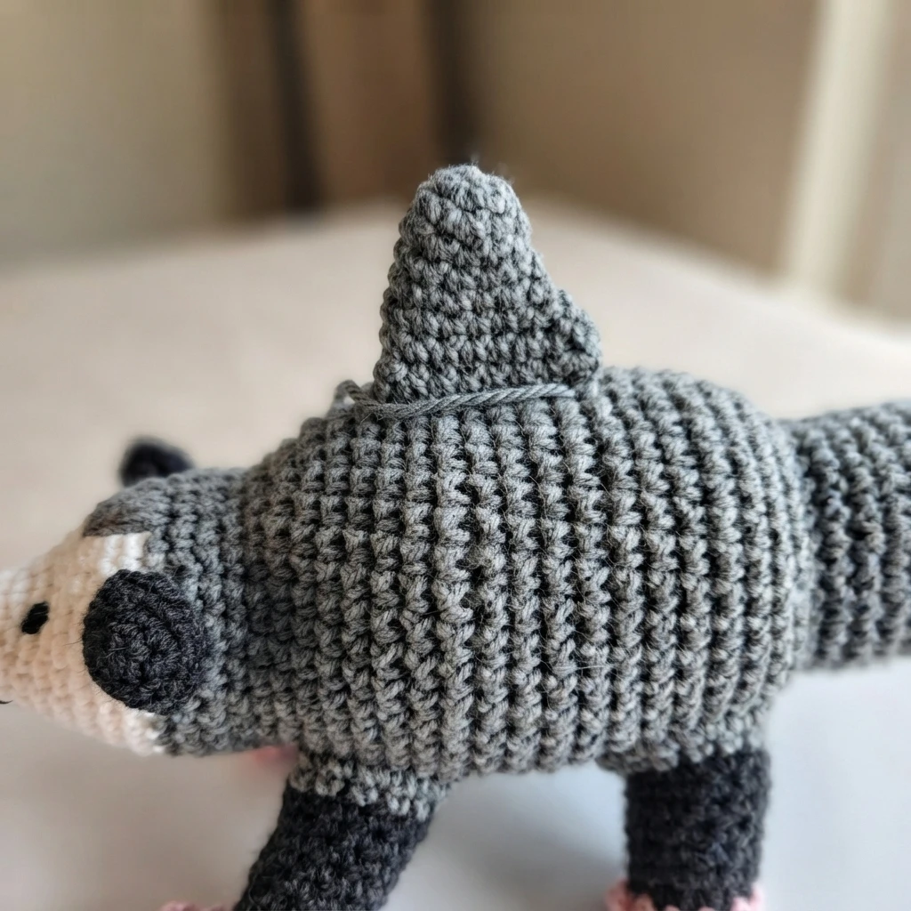 Close-up of a gray crocheted shark fin attached to the middle of a gray amigurumi body, with a black ear, cream muzzle and a loose strand of gray yarn lying across the back
