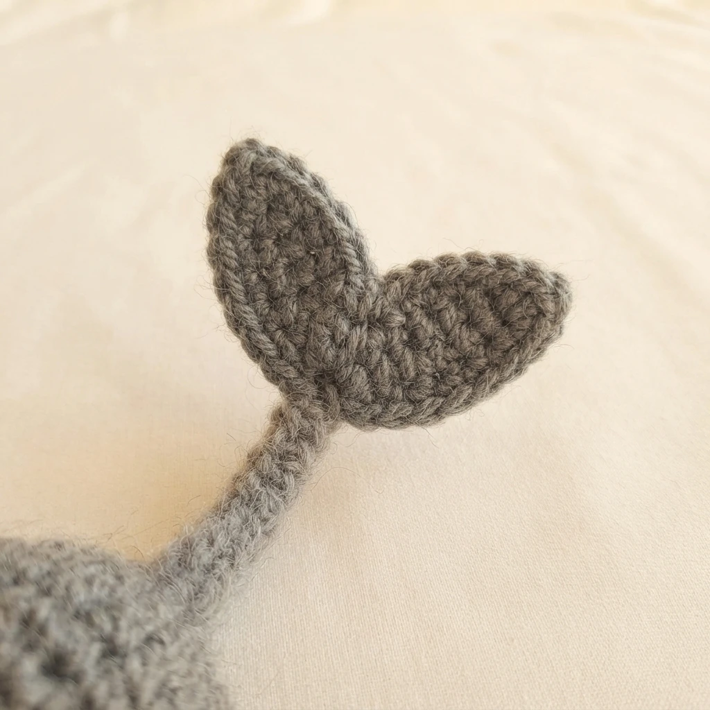 Two small gray crocheted fins sewn together at their bases to form a V shaped tail fin, attached to a gray crocheted tail, resting on a light cream fabric surface.