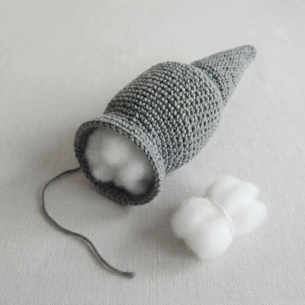 Gray crocheted cone-shaped tail piece with an open end showing white stuffing inside, a long loose gray yarn strand, and a clump of polyester stuffing resting beside it on a light fabric surface.