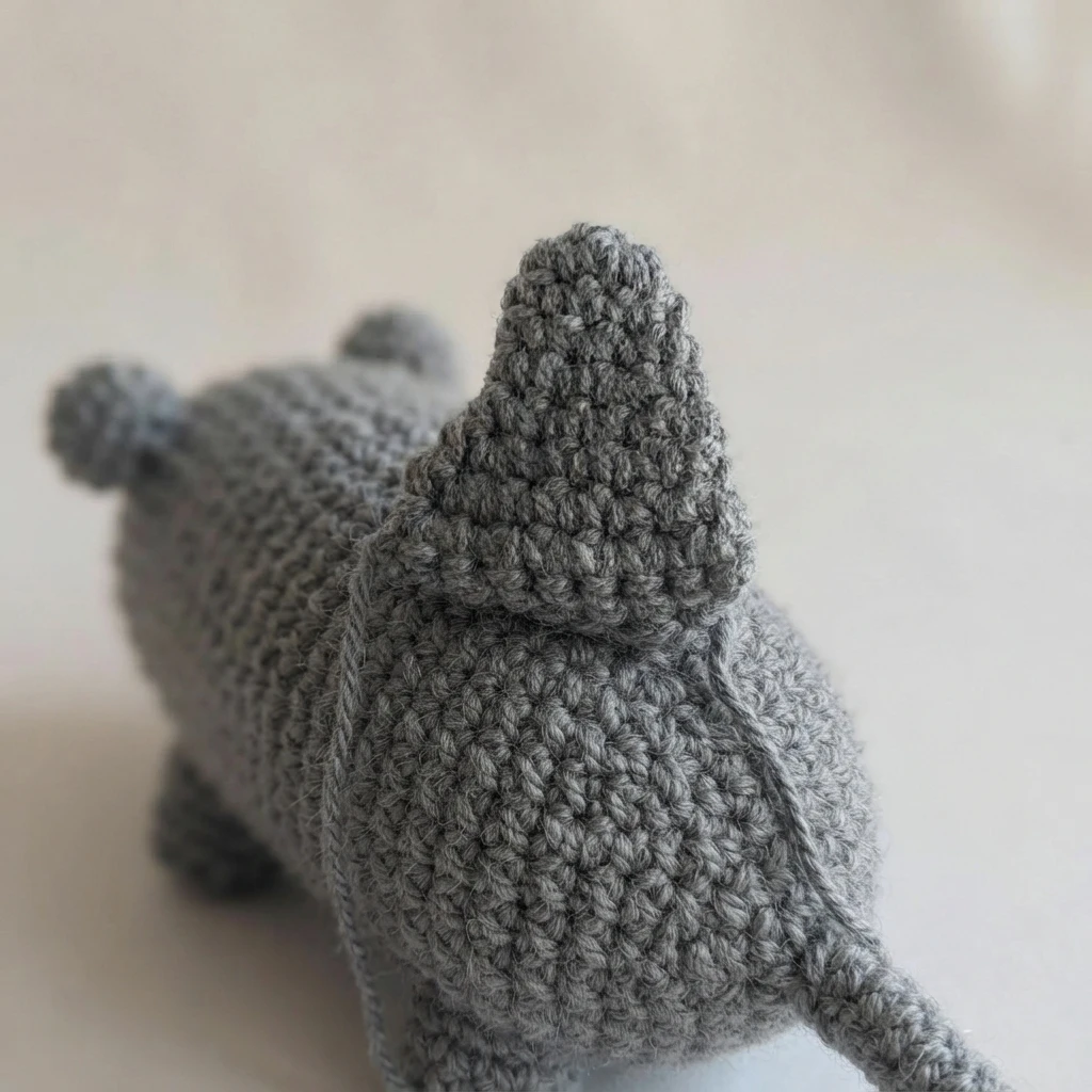 Close-up of a gray crocheted plush seen from behind, showing a triangular knitted fin sewn onto the middle of its back, with small rounded ears and part of the body and legs visible against a plain light background.