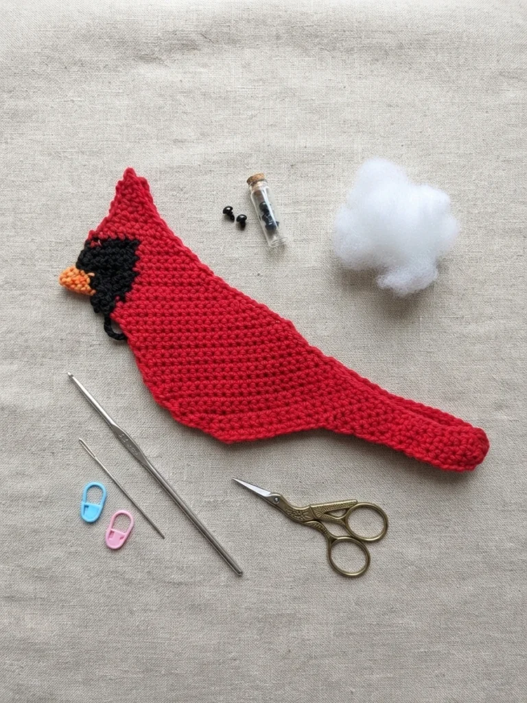 Flat lay of a red crochet cardinal front panel with a black face mask and orange beak, lying on linen fabric with a crochet hook, a tapestry needle, two stitch markers, small metal scissors, two black safety eyes in a glass vial and a pile